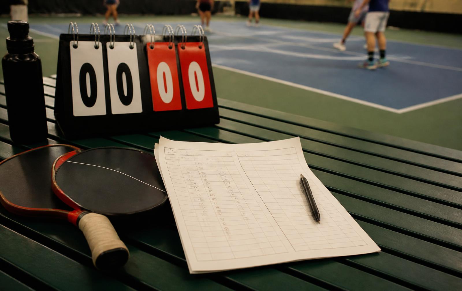 Courtside flip scoreboard and a handwritten score sheet beside paddles